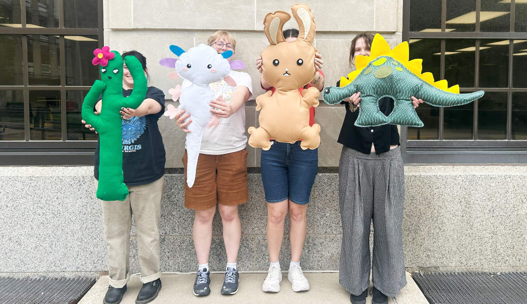 four students holding large felt stuffies outside the Memorial Union. From left to right they're holding a green cactus, a white axolotl, a tan bunny, and a green dinosaur